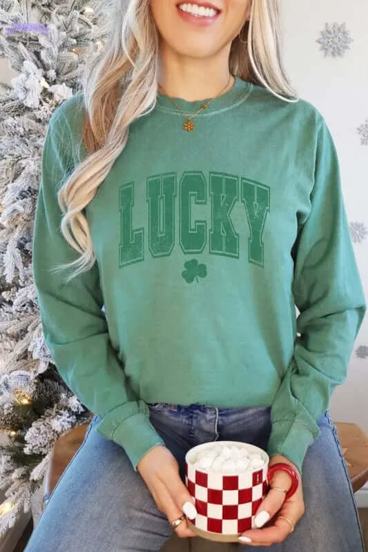 Woman wearing a green long sleeve shirt with "LUCKY" graphic, holding a red and white checkered mug with hot chocolate. Aaria Tees