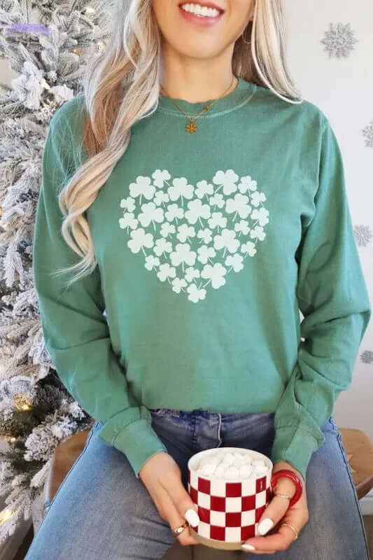 Woman wearing Shamrock Heart St Patrick's Day long sleeve tee, holding a cup with marshmallows, festive backdrop.