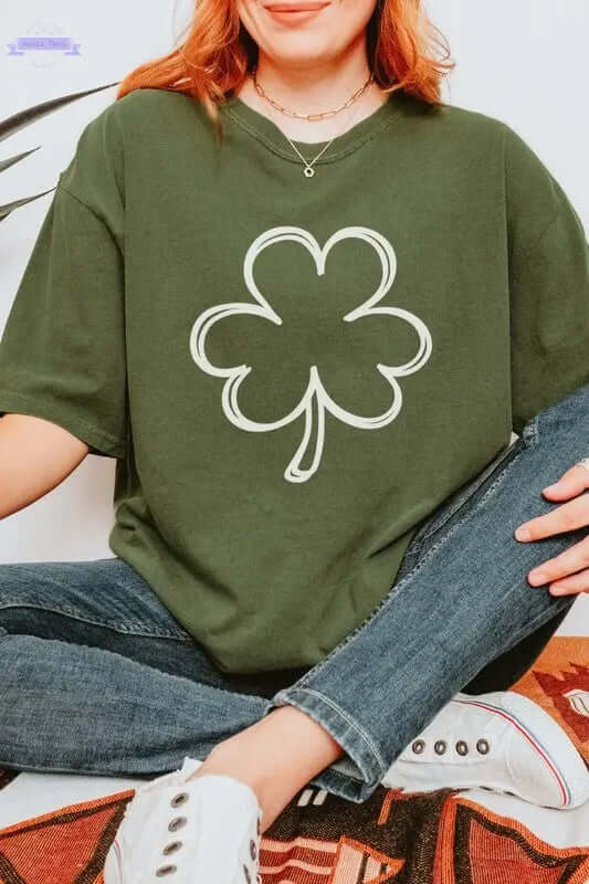 Woman wearing a green long sleeve shirt with a white shamrock graphic, sitting on a patterned surface.